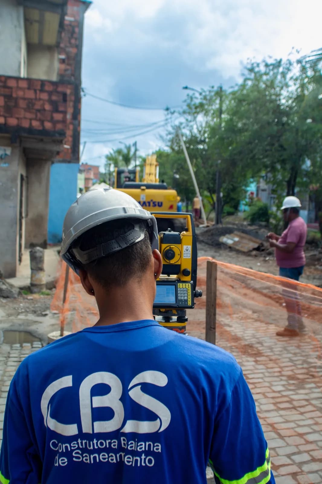 Retroescavadeira JCB executando escavação em rua urbana durante obra de infraestrutura, com colaboradores sinalizando o entorno